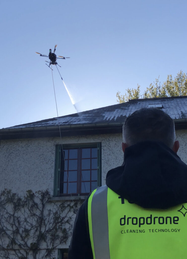 Drone en cours de nettoyage d'une toiture de maison individuelle sous la supervision d'un technicien Dropdrone